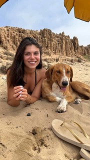 Young smiling woman with dark hair lying on a sandy beach next to a happy brown dog, both relaxing