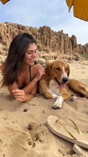 Woman lying on sand, smiling and gently petting a dog, relaxing on a beach with cliff formations in