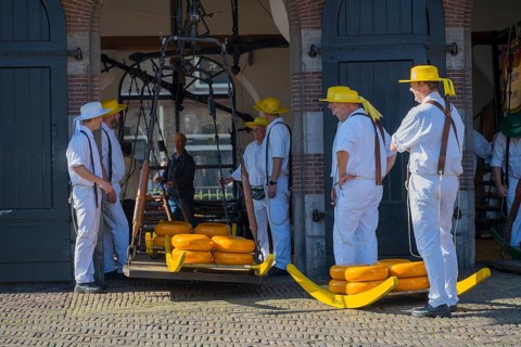 Männer mit gelben Hüten und weißer Kleidung tragen Käse auf einem Tragegestell zur ehemaligen