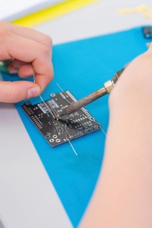 Person soldering an electronic circuit board with a soldering iron on a blue mat