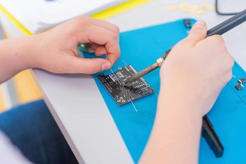 A person solders a circuit board with a soldering iron, surrounded by electronic parts