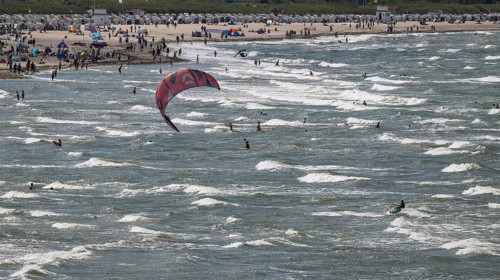 Kitesurfers and holidaymakers on the beach and in the sea, waves, beach chairs in the background,
