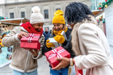 Three happy friends opening christmas gifts at a christmas market, enjoying winter holidays