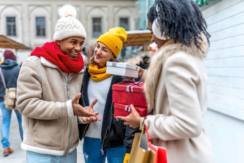 Three friends exchanging christmas gifts and shopping bags at a winter market, enjoying the festive