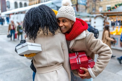 Two friends joyfully exchanging gifts at a bustling outdoor christmas market, surrounded by festive