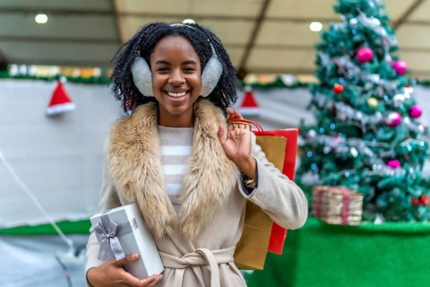 Young black woman wearing earmuffs holding christmas present and shopping bags, smiling in front of