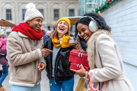Three friends are having fun exchanging christmas gifts at a winter market, laughing and enjoying