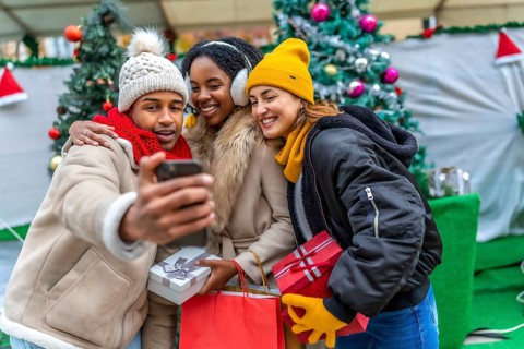 Three happy friends taking a selfie with smartphone at the christmas market, holding gifts and