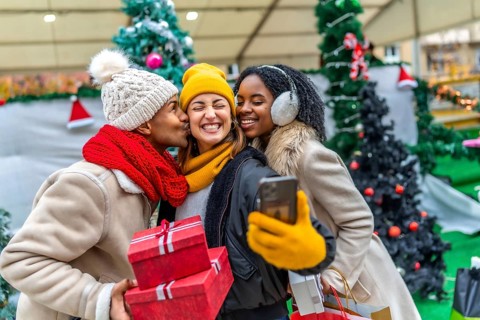 Three happy friends are taking a selfie at a christmas market, one of them is kissing another one