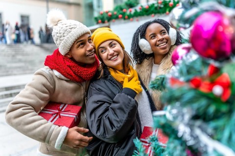 Three friends holding christmas gifts are admiring a decorated christmas tree at a city market