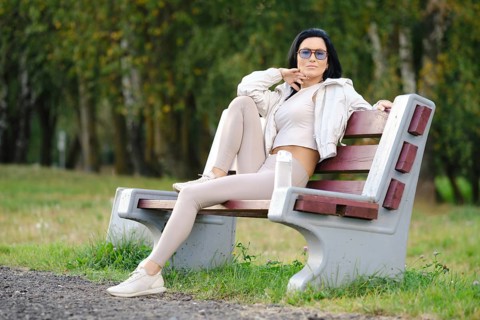 A woman sits on a park bench dressed in light-colored casual clothes, sunglasses on, with a relaxed
