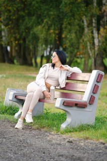 A woman dressed in comfortable casual wear sits on a park bench, gazing thoughtfully into the