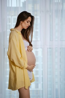 A pregnant woman stands near a window in profile, wearing a yellow shirt and white outfit, lovingly
