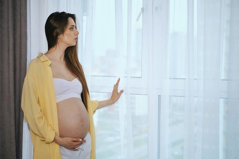 A pregnant woman stands by a window in a contemporary apartment, wearing a white top and loose