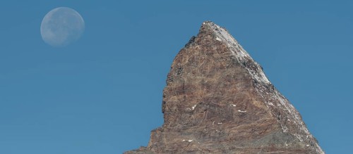 The Matterhorn mountain peak rises steeply against a bright blue sky. The full moon glistens gently