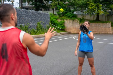 Two baseball players practicing catching on a court, with a woman wearing a glove attempting to