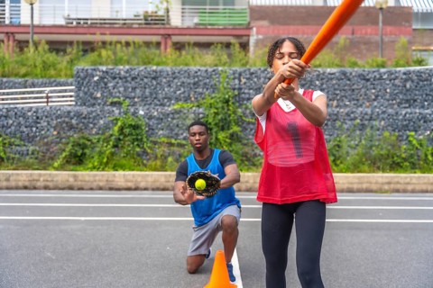 Engaging in an outdoor baseball practice, a woman swings a bat with focus while a man kneels,