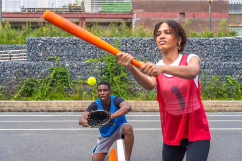 Female baseball player swinging a bat to hit a ball while a male catcher prepares to catch it.