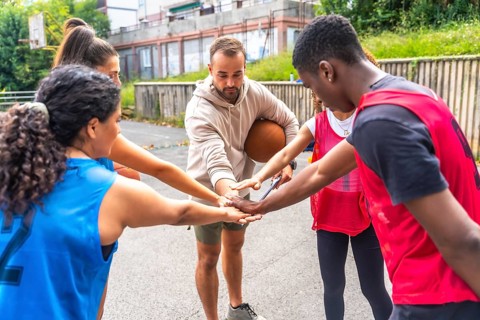 Diverse group of basketball players and their coach gather in a huddle, joining hands together,