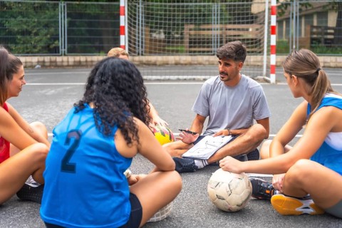 Soccer coach sitting on the ground, using a notebook to explain game strategies to female athletes