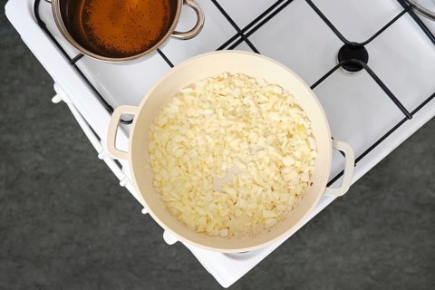 Chopped onions cooking in a white pan over medium heat on a stove. A pot of simmering broth sits