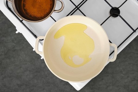 Cooking oil is poured into a frying pan on a stovetop. A pot with vegetable stock sits nearby,