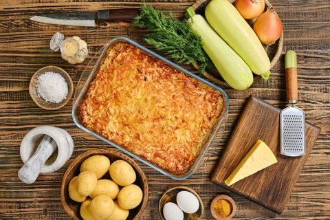 Loaded baked potato casserole sits in a glass dish surrounded by fresh vegetables and spices.