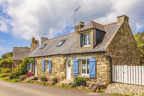 Old idyllic stone cottage with blue shutters and blooming flowers at the house wall by a road in