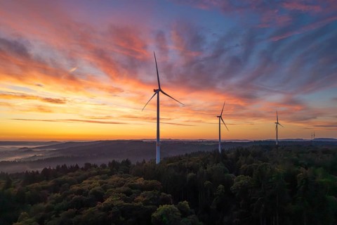 Three wind turbines in the foreground of a colourful morning sky over a hilly landscape, near