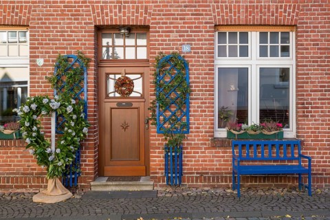 A decorated brick house façade with a blue bench and floral decorations next to the entrance, North