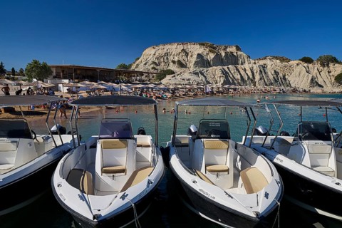 Several boats are moored on a jetty with a rocky beach in the background. Holidaymakers relaxing on