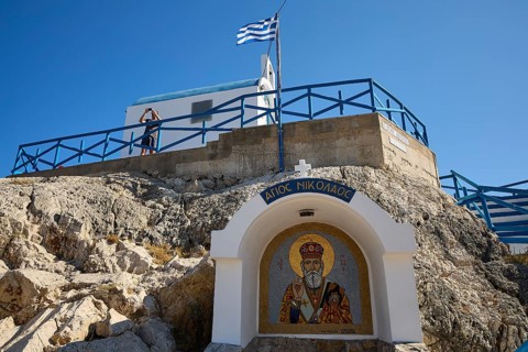 Chapel in rock formation with icon and Greek flag in the foreground, Chapel of Agios Nikolaos,