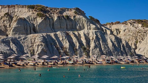 Beach in front of a large rock face, lined with sun loungers and umbrellas, bathers enjoying the