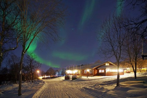 An illuminated house under auroras with snow-covered trees on a winter night, Sortland campsite,