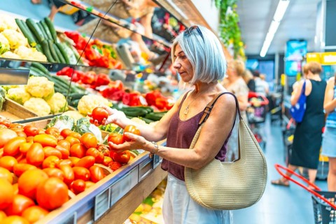 Mature woman with gray hair choosing ripe tomatoes in a grocery store, surrounded by colorful