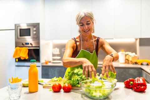 Elderly woman wearing an apron, making a salad with fresh ingredients in a bright kitchen,