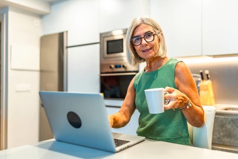 Smiling senior freelancer woman enjoying a cup of coffee while working on her laptop in her modern