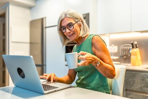 Smiling senior woman using laptop and drinking coffee in modern kitchen, enjoying work from home or