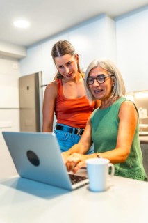 Senior woman using a laptop with guidance from a young woman in a modern kitchen, sharing knowledge