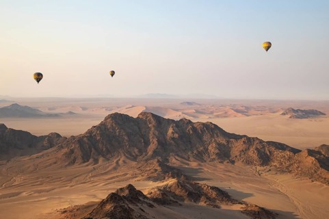 Three hot-air balloons above an arid plain and isolated mountain ridges at the edge of the Namib