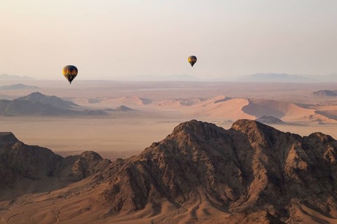 Two hot-air balloons above an arid plain and isolated mountain ridges at the edge of the Namib