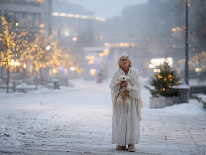 An elderly woman in a nightgown stands with her teddy bear in a cold, wintry environment, looking