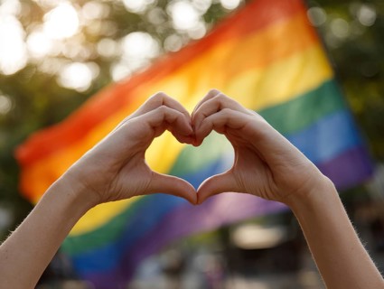 Two hands form a heart in front of a waving rainbow flag in the sunlight, symbolising equality,