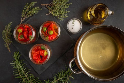 Glasses filled with chilli peppers next to a saucepan with vinegar, oil and herbs on a dark