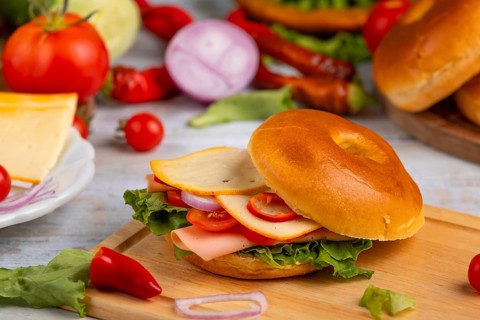 A bagel topped with cheese, sausage, tomatoes, onion and lettuce leaves on a wooden board