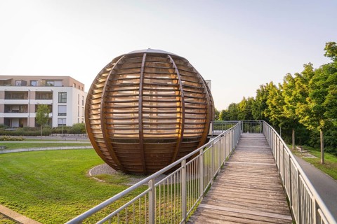 Wooden spherical structure in front of modern residential building at sunset in urban park, small