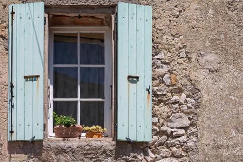 A window with blue shutters in a rustic house wall with flowering potted plants, Provence, France
