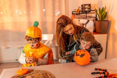 Mother and children wearing halloween costumes are sitting at a table at home, playing with pumpkin
