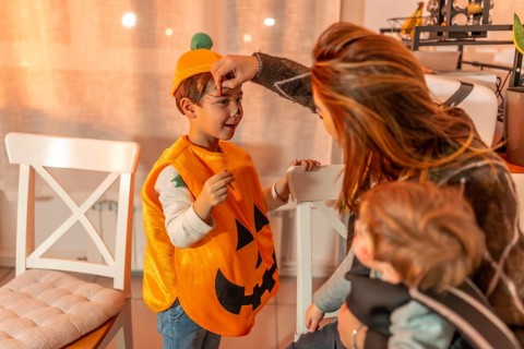 Mother applying halloween makeup on her son wearing a pumpkin costume, while holding her baby