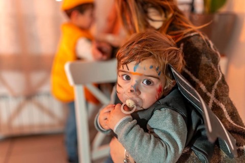 Toddler wearing a halloween costume with painted face and pacifier, held by mother, with another